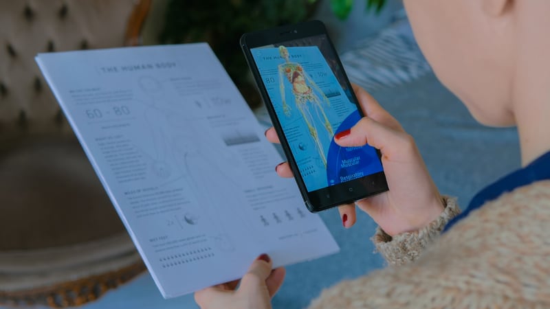 An Over-the-shoulder Shot Of A Woman Holding A Health Document In Her Left Hand, While Her Smartphone Is In The Right. The Document Is In Black And White, And The Phone Shows An In-color Version - Indicating A Pop-out Version The Text Is Presumably Referring To, Signifying Augmented Reality.