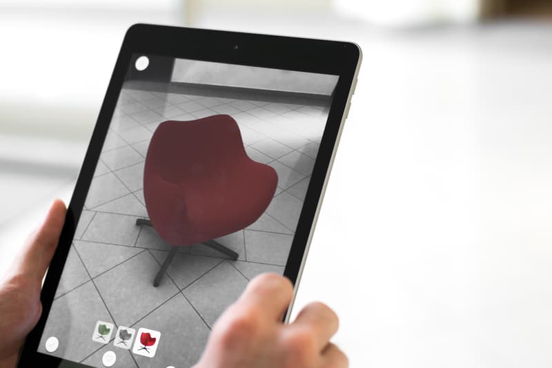 A Picture Showcasing Augmented Reality. A Pair Of Hands Are Holding A Tablet, With A Plain Floor In The Background. However, On The Tablet, It Shows A Modern Chair In A Burgundy/red Color.