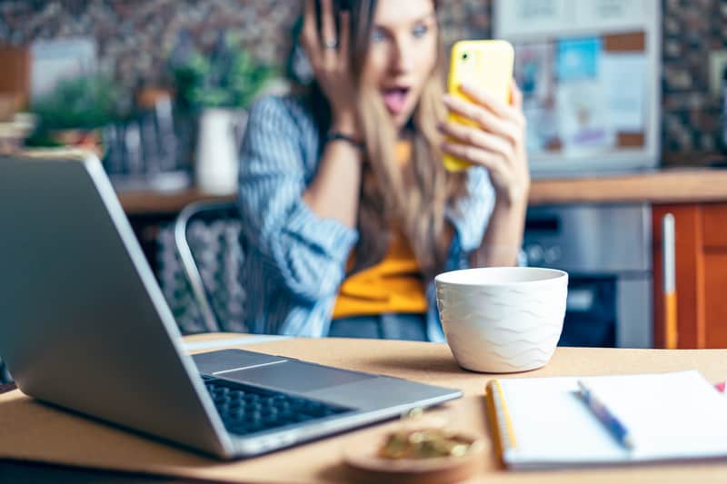 Woman Holding Up Yellow Phone In Her Left Hand. The Other Hand Is On The Side Of Her Face And She Is Making A Shocked, Excited Expression