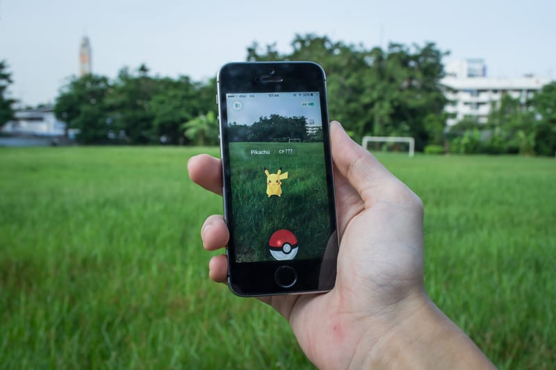 There Is A Hand Holding A Phone, Playing Pokémon GO With Pikachu In The Center Of The App.